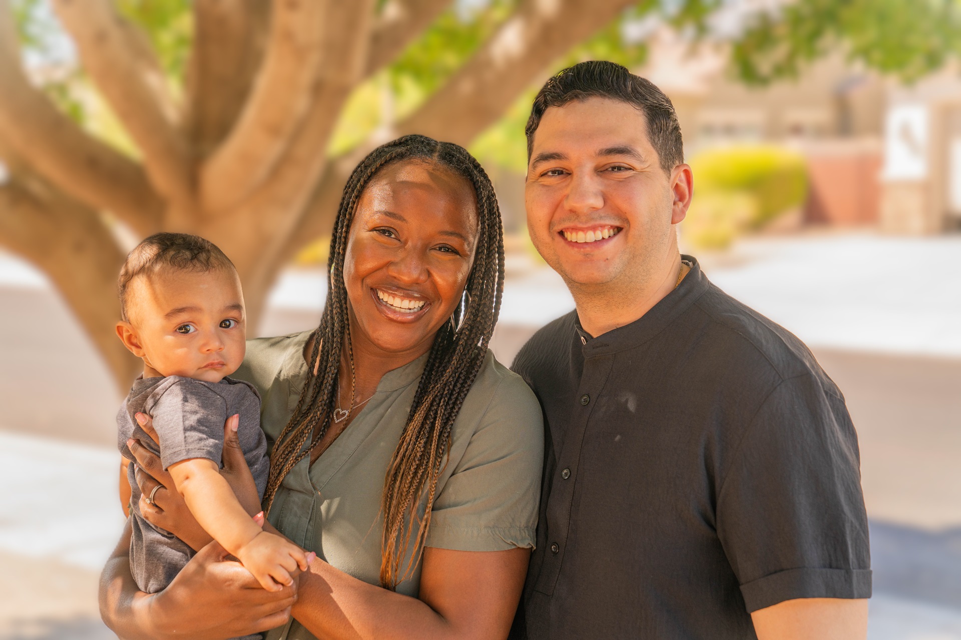 Family of three smiling outdoors under a tree in natural sunlight