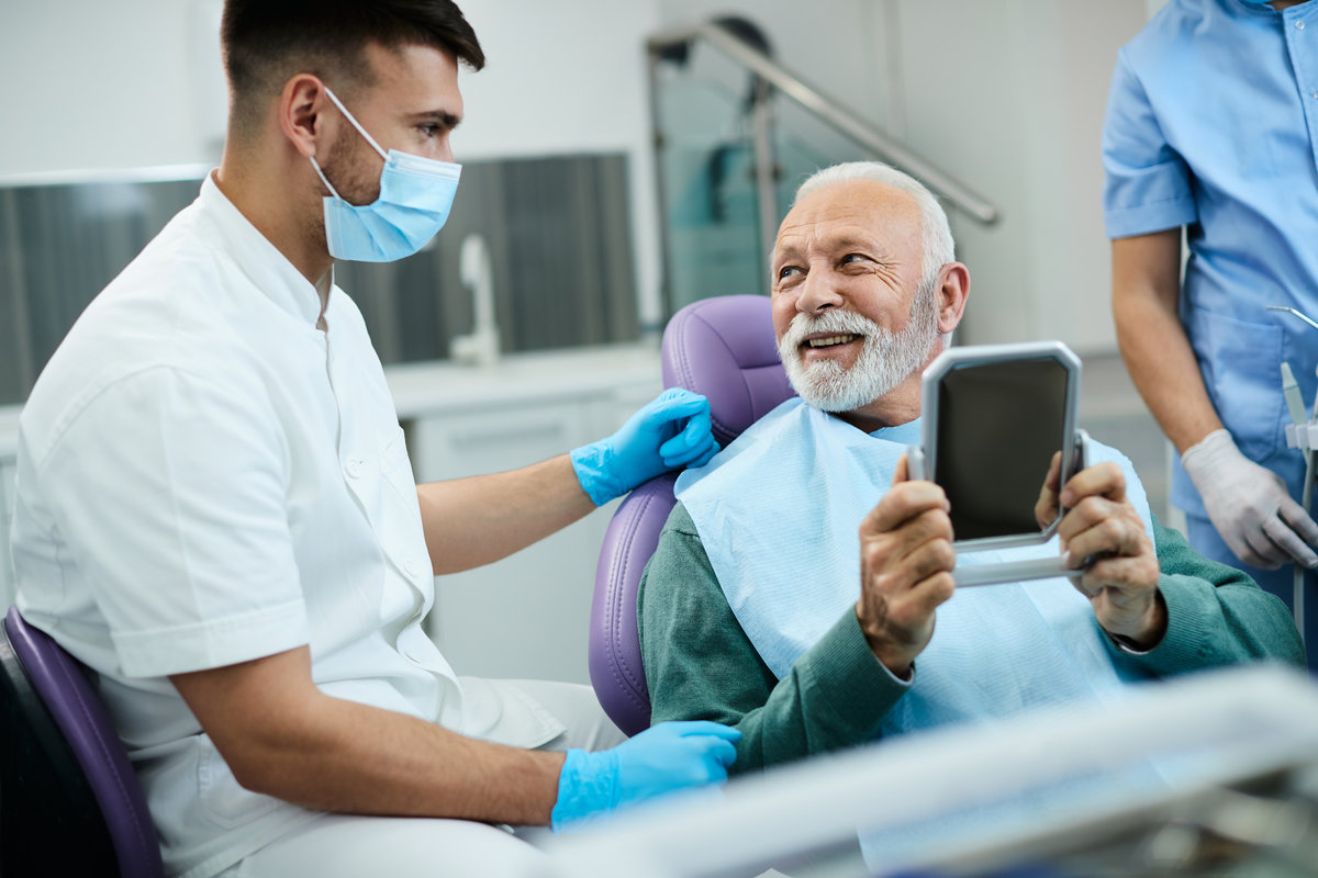 Satisfied senior man talks with dentist while looking his teeth in mirror at dentist's office.