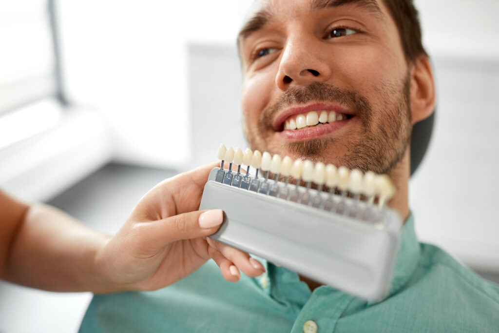 medicine, dentistry and healthcare concept - female dentist with tooth color samples choosing shade for male patient teeth at dental clinic